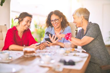 Orta yaşlı kadınlarla buluşup, öğle yemeği yiyip, kahve içiyorlar. Olgun arkadaşlar mutlu bir şekilde gülümsüyor. Güneşli bir günde evde akıllı telefon kullanıyorlar.