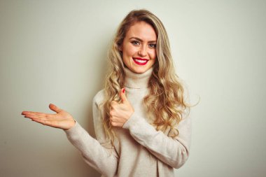 Kış balıkçı yaka kazak giyen güzel bir kadın izole edilmiş beyaz arka planda avuç içini gösteriyor ve baş parmaklarıyla hareket ediyor, mutlu ve neşeli gülümsüyor.