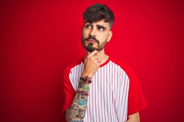Çizgili tişörtü olan genç bir adam izole edilmiş kırmızı arka plan üzerinde duruyor ve elleri çenesinde soru sormayı düşünüyor, düşünceli bir ifade. Düşünceli bir yüzle gülümsüyor. Şüphe kavramı.