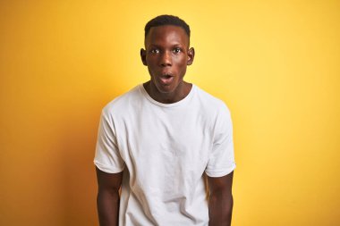Beyaz t-shirt giyen genç Afro-Amerikalı adam izole edilmiş sarı arka planda duruyor. Şaşırtıcı bir ifade, korku ve heyecanlı bir yüz ifadesi var..