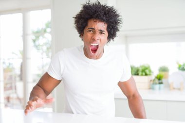 Genç afro-amerikan adam evde oturan rahat beyaz t-shirt giyen kızgın ve deli çığlık sinirli ve öfkeli, öfke ile bağırıyor. Öfke ve agresif kavram.