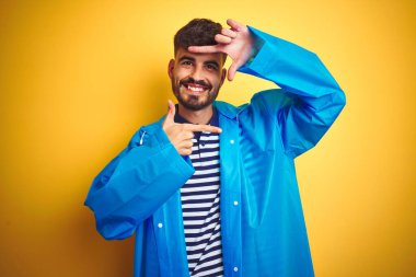 Yağmurluk giyen genç yakışıklı adam izole edilmiş sarı arka planda duruyor gülümsüyor ve elleri ve parmakları gülen yüzlü bir çerçeve yapıyor. Yaratıcılık ve fotoğrafçılık kavramı.