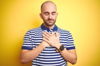Sakallı, sarı izole edilmiş arka plan üzerine sıradan mavi tişört giyen, elleri göğsünde, gözleri kapalı ve yüzünde minnet dolu bir jest olan genç kel bir adam. Sağlık kavramı.