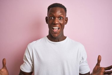 Beyaz tişört giyen Afro-Amerikalı adam pembe arka planda mutlu ve heyecanlı duruyor. Kazanan ifade zaferi büyük bir gülümsemeyle kutluyor ve ellerini kaldırıyor.