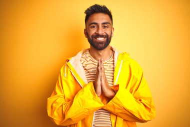 Yağmurluk giyen genç Hintli adam izole edilmiş sarı arka planda duruyor ellerle dua ediyor birlikte af diliyor kendinden emin bir şekilde gülümsüyor..