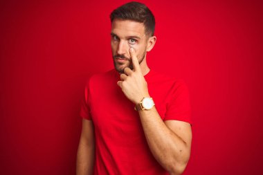 Kırmızı izole edilmiş arka plan üzerine gündelik tişört giyen genç yakışıklı adam senin jestini, şüpheli ifadesini izliyor.