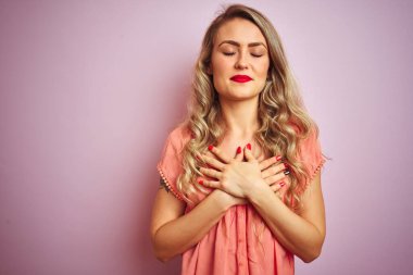 Pembe izole edilmiş arka planda tişört giyen genç ve güzel bir kadın. Elleri göğsünde, gözleri kapalı ve yüzünde minnet dolu bir jest ile gülümsüyor. Sağlık kavramı.