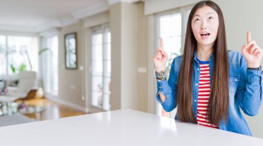 Uzun saçlı, güzel Asyalı kadın kot ceket giyiyor. Şaşırmış, şaşırmış, parmaklarıyla işaret ediyor ve kollarını kaldırıyor..