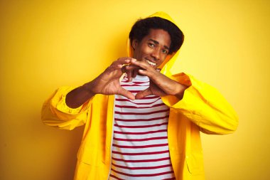 Siyah saçlı, yağmurluk giyen, izole edilmiş sarı arka planda duran, kalp sembolü ve elleri ile şekil veren Afrikalı bir adam. Romantik konsept.