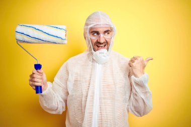 Ressam kıyafeti giyen ve elinde boya rulosu olan genç bir adam izole edilmiş sarı arka plan üzerinde mutlu bir gülümsemeyle baş parmağıyla yüzünü gösteriyor.