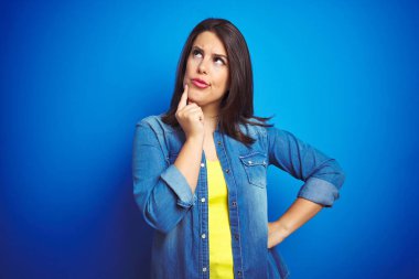 Genç, güzel, esmer kadın mavi kot ceket giyiyor ve mavi izole edilmiş arka plan üzerinde elleri çenesinde soru sormayı, düşünceli yüz ifadesini düşünüyor. Düşünceli bir yüzle gülümsüyor. Şüphe kavramı.