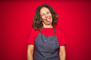 Kırmızı izole arka planda önlük giyen orta yaşlı bir kadın komik bir ifadeyle dilini dışarı çıkartıyor. Duygu kavramı.