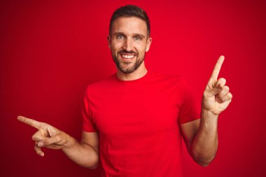Kırmızı izole edilmiş arka plan üzerine gündelik tişört giyen genç yakışıklı adam farklı yönleri gösteren kendine güvenle gülümsüyor. Reklam için boşluk kopyala