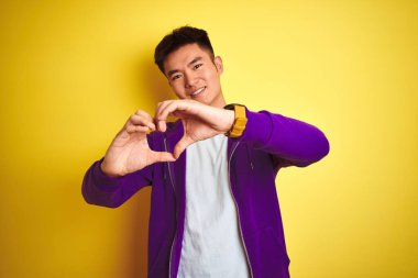 Mor kazak giyen Asyalı genç adam izole edilmiş sarı arka planda duruyor. Aşkla gülümsüyor, elleriyle kalp sembolü yapıyor. Romantik konsept.