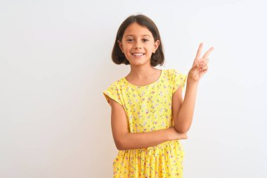 Sarı çiçekli elbise giyen güzel bir kız çocuğu izole edilmiş beyaz arka planda dikiliyor ve kameraya gülümsüyor ve parmaklarıyla zafer işareti yapıyor. İki numara..