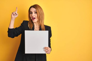 Temiz gözlü genç kızıl iş kadını izole edilmiş sarı arka planda afiş tutarken bir fikir ya da soru işareti ile şaşırdı.