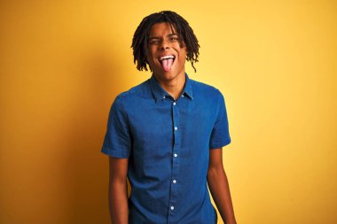 Rastgele kot gömlek giyen Afro bir adam izole edilmiş sarı arka planda dikiliyor ve komik bir ifadeyle dilini dışarı çıkarıyor. Duygu kavramı.