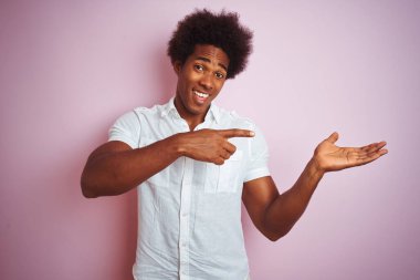 Afro saçlı, beyaz tişörtlü genç bir adam izole edilmiş pembe arka planda durup kameraya hayret ediyor ve gülümsüyor..