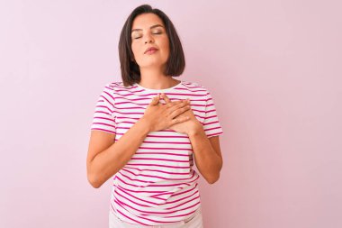 İzole edilmiş pembe arka plan üzerinde duran çizgili tişört giyen genç ve güzel bir kadın. Elleri göğsünde, gözleri kapalı ve yüzünde minnet dolu bir jest ile gülümsüyor. Sağlık kavramı.
