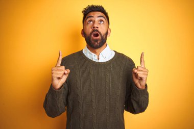 Yeşil süveter ve tişört giyen genç Hintli adam sarı arka plan üzerinde duruyordu. Şaşırmış ve şaşırmıştı. Parmaklarıyla işaret ediyor ve kollarını kaldırıyordu..