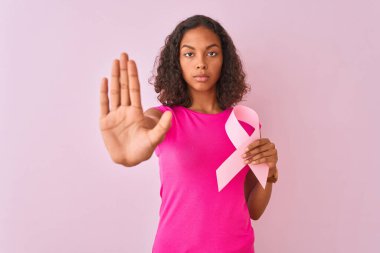 Kanser kurdelesi tutan genç Brezilyalı kadın pembe arka planda duruyor. Açık elle dur işareti yapıyor. Ciddi ve kendinden emin bir ifadeyle, savunma hareketi yapıyor.