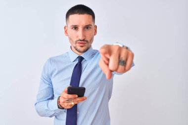 Genç yakışıklı iş adamı, izole edilmiş arka plan üzerinde akıllı telefon kullanıyor. Kamerayı işaret ediyor ve sen, el işareti, ön taraftan pozitif ve kendinden emin bir hareket.