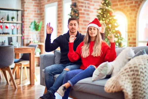 一对年轻的夫妇 头戴圣爪帽 坐在沙发上 围坐在圣诞树旁 微笑着 闭上双眼 用手指做冥想手势 瑜伽概念 — 图库照片