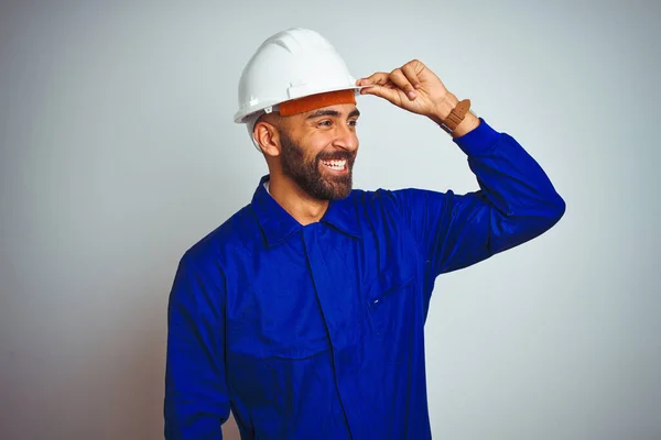 Handsome indian worker man wearing uniform and helmet over isolated white background very happy and smiling looking far away with hand over head. Searching concept.