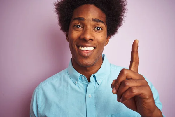 Young american man with afro hair wearing blue shirt standing over isolated pink background surprised with an idea or question pointing finger with happy face, number one