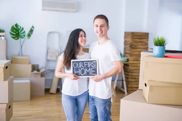 Young beautiful couple standing on the floor holding blackboard with message at new home around cardboard boxes