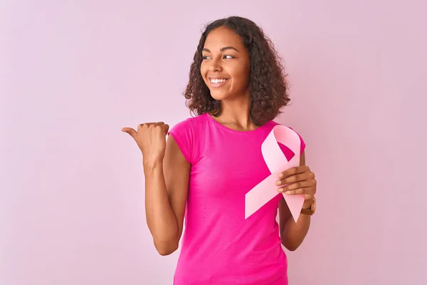 若いですブラジル人女性保持癌リボン立っています上の隔離されたピンクの背景指すと表示しますとともに親指で側まで幸せな顔笑顔 — ストック写真