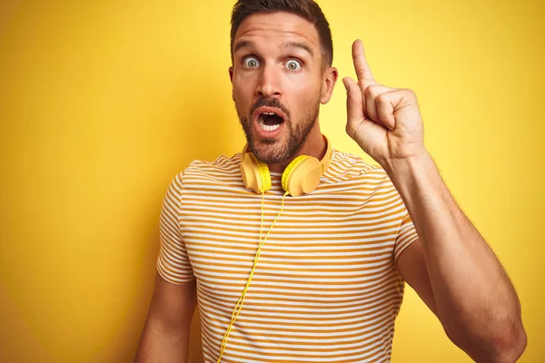 黄色の隔離された背景の上の音楽を聞くヘッドフォンを身に着けている若いハンサムな人は巧妙な考えと指を指す 興奮し 第一番 — ストック写真