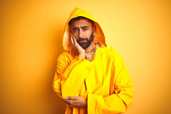 Young indian man wearing raincoat with hood standing over isolated yellow background thinking looking tired and bored with depression problems with crossed arms.