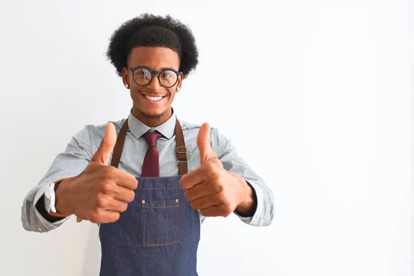若いアフリカ系アメリカ人の店主は 孤立した白い背景の上にエプロンの眼鏡をかけて 手で積極的なジェスチャーをすることを承認し 親指を上げて笑顔と成功に満足しています 勝者のジェスチャー — ストック写真
