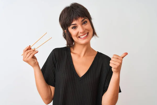 年轻美丽的女人拿着筷子站在孤立的白色背景上 用拇指指向旁边 笑容满面 — 图库照片