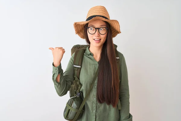 Chinese hiker woman wearing canteen hat glasses backpack over isolated white background smiling with happy face looking and pointing to the side with thumb up.