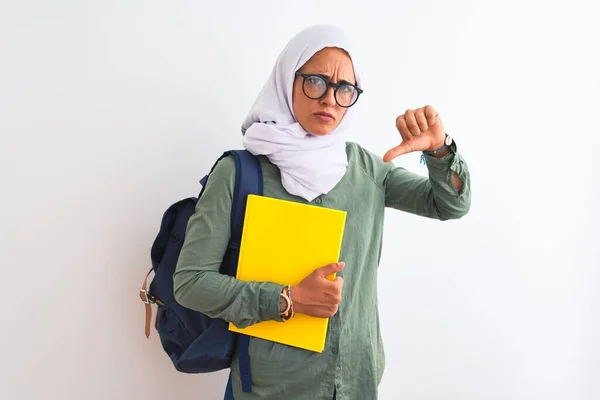 Young Arab student woman wearing hijab and backpack holding a book over isolated background with angry face, negative sign showing dislike with thumbs down, rejection concept