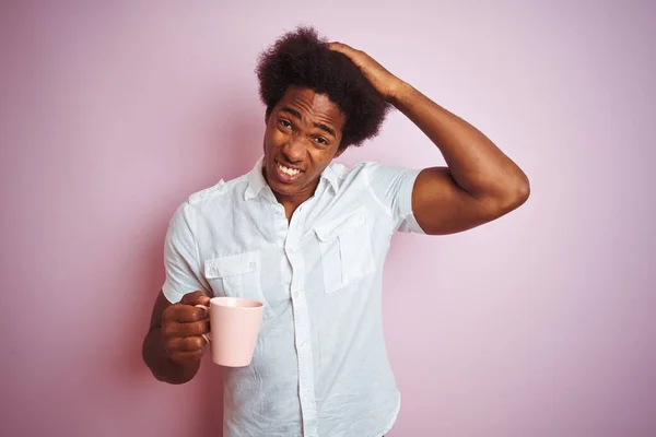 Young african american man drinking a cup of coffee standing over isolated pink background stressed with hand on head, shocked with shame and surprise face, angry and frustrated. Fear and upset for mistake.