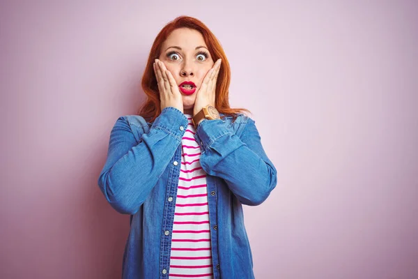 Beautiful redhead woman wearing denim shirt and striped t-shirt over isolated pink background afraid and shocked, surprise and amazed expression with hands on face