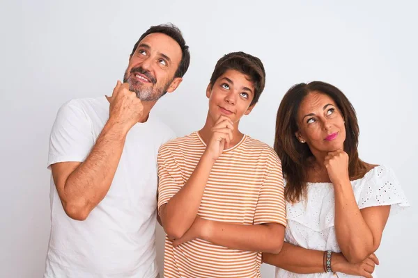 Family of three, mother, father and son standing over white isolated background with hand on chin thinking about question, pensive expression. Smiling with thoughtful face. Doubt concept.