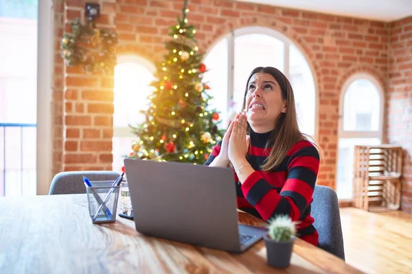 Beautiful woman sitting at the table working with laptop at home around christmas tree begging and praying with hands together with hope expression on face very emotional and worried. Asking for forgiveness. Religion concept.