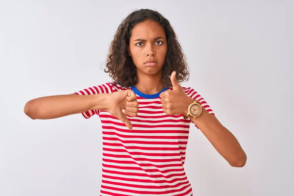 隔離された白い背景の上に立つ赤い縞模様のTシャツを着た若いブラジル人女性親指を上下に 意見の相違および一致の表現をする 狂気の葛藤 — ストック写真