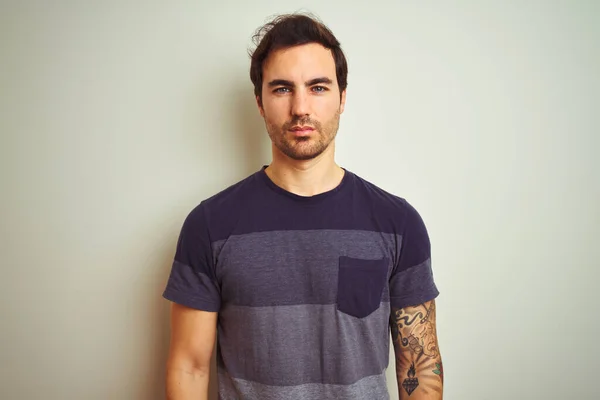 Young handsome man with tattoo wearing casual t-shirt over isolated white background with serious expression on face. Simple and natural looking at the camera.