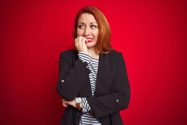 Beautiful redhead business woman wearing elegant jacket over isolated red background looking stressed and nervous with hands on mouth biting nails. Anxiety problem.