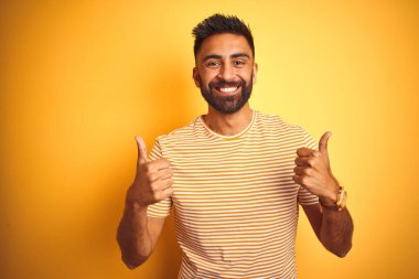 İzole edilmiş sarı arka plan başarısı tabelasının üzerinde duran genç Hintli adam el hareketleriyle olumlu bir hareket yapıyor, gülümsüyor ve mutlu. Neşeli ifade ve kazanan jest.