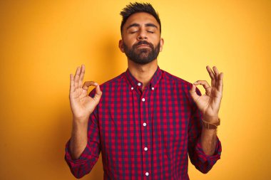 İzole edilmiş sarı arka plan üzerinde duran sıradan bir gömlek giyen genç Hintli adam rahatla ve parmaklarıyla meditasyon hareketi yaparken gözleri kapalı gülümse. Yoga konsepti.