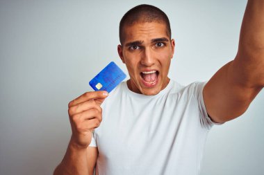 Genç ve yakışıklı bir adam kredi kartını izole edilmiş beyaz bir arka planda tutarken sinirlenmiş ve sinirlenmiş bir şekilde bağırıyor, çıldırmış ve bağırıyor.