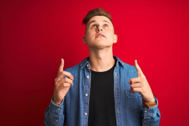 Soyutlanmış kırmızı arka plan üzerinde duran kot gömlek giyen genç yakışıklı adam şaşırmış ve şaşırmış bir şekilde yukarı bakıyor ve parmaklarıyla işaret ediyor ve kollarını kaldırıyordu..