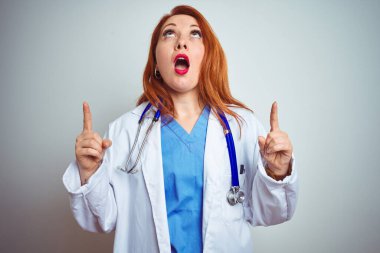 Genç kızıl saçlı kadın steteskop kullanıyor. İzole edilmiş beyaz bir zemin üzerinde. Şaşırmış ve şaşırmış..