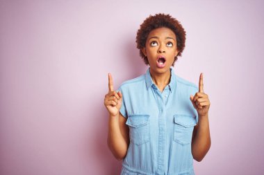 Afro saçlı, güzel, Afro-Amerikalı bir kadın. Pembe arka planı var. Şaşırmış ve şaşırmış bir şekilde yukarı bakıyor ve parmaklarıyla işaret ediyor..